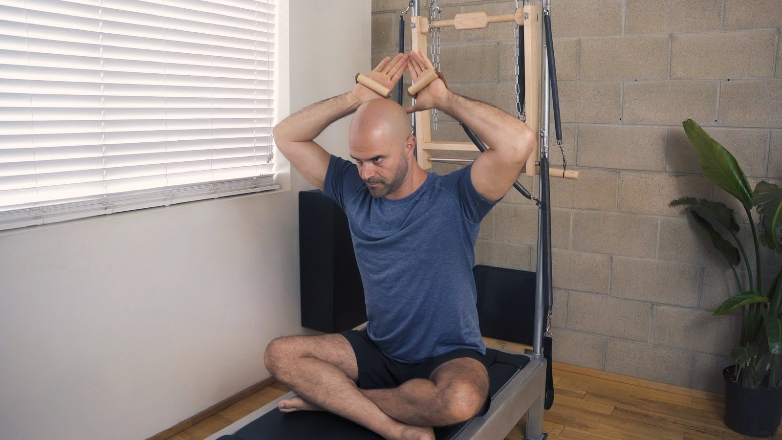 Yonatan seated cross-legged on the reformer, holding the tower handles overhead, in the West Hollywood Pilates studio.
