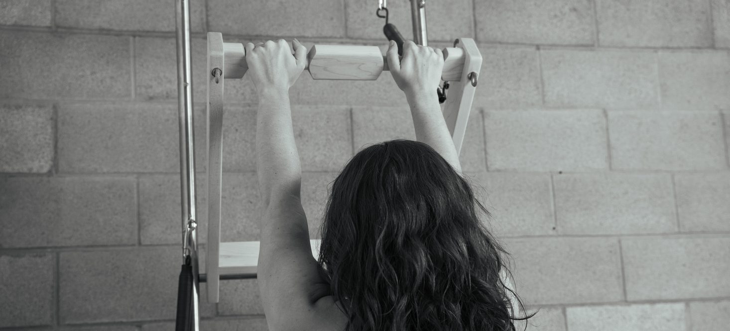 A client hanging from the Cadillac push-through bar, both hands gripping the wooden bar, seen from behind against the studio's block wall
